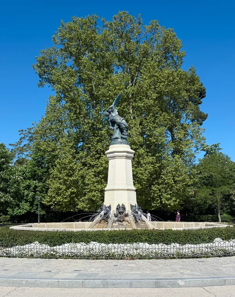 The Fuente del Ángel Caído seen frontally, with the sculpture of Lucifer on the pedestal and a large tree behind it, in Parque del Retiro, Madrid.