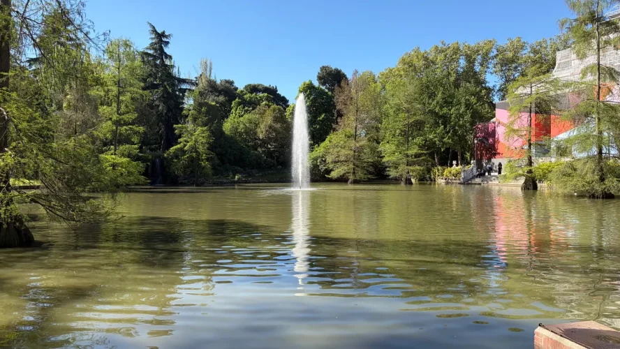 The pond by the Palacio de Cristal in Parque del Retiro, Madrid, surrounded by trees, with the building visible under restoration on the right.