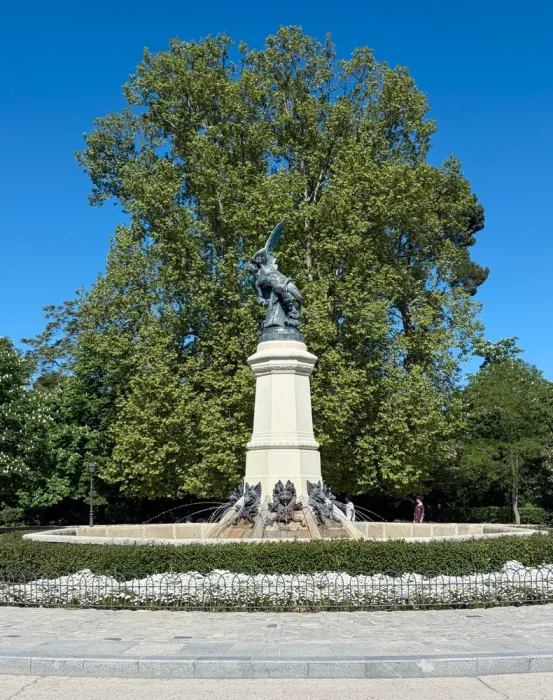 The Fuente del Ángel Caído seen frontally, with the sculpture of Lucifer on the pedestal and a large tree behind it, in Parque del Retiro, Madrid.